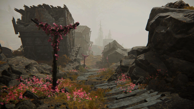 A view down the stairs, back into the village. There's a profusion of pink flowers around a cruciform post in the foreground, and a large grey outcrop of rock on the other side of the path. The houses are tumbledown, their stonework broken, their timbers jagged.