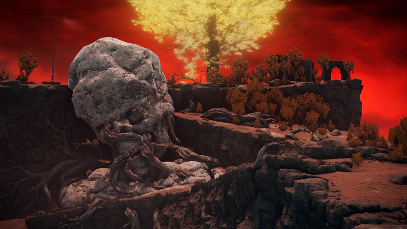 A winding path up a red-brown landscape. A vast stone skull, pitted with coral-like lesions, rests against the hillside, twisted tree-branches crawling through its eyes and mouth. Ruined stone structures line the clifftops, beneath the light of a minor erdtree.