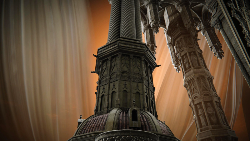 A close-up of a large grey-stone column atop a red-tiled domed building. The lower part is octagonal and ornately fashioned into arches and symbols, with horned motifs; the upper part becomes three round pillars with spiralling patterns. To the right is another column with multiple layers, each containing a row of carved humanoid figures, and above that a cluster of dangling columns from the stonework above, each terminating in a blunt point. The sky is full of veils cast orange by the sunsert.