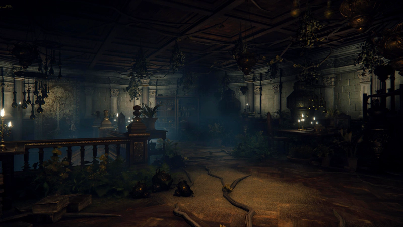A dimly-lit chamber with roots snaking across the floor and assorted plantlife hanging from the ceiling and growing from the floor. Amidst the foliage, there are tables and chairs, bookshelves and large jars.