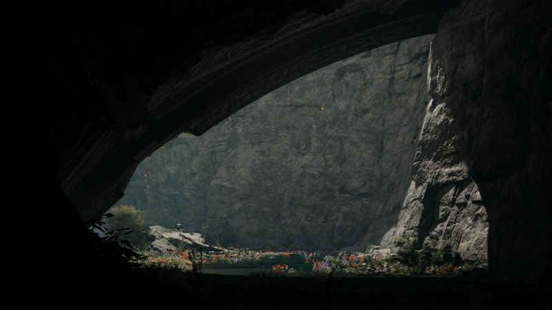 A view through an arched ruin to where sunlight drenches a shallow river, speckled with red and blue wildflowers. To the right, the cliffside is grey and crisp in the sunshine, and in the distance a corpse sits cross-legged on a rock.