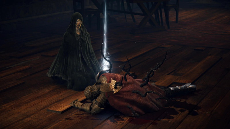 Fia, a woman in black robes, kneels over the body of D, a warrior in gold and silver armour with a red cloak. Black vines are growing from his back, and blood pools behind him. We can partially see below Fia's hood, and her expression is not one of distress.