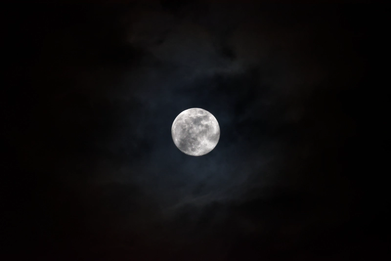 A bright full moon glowing through layered clouds. The moonshine makes the clouds glow grey-blue, surrounded by an indistinct copper-coloured ring of lunar corona.