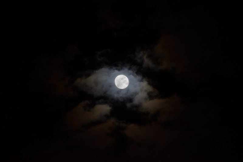 A bright full moon shining through a lozenge-shaped cloud. Other small clouds hover around the edges of the image, cast red by the lunar corona.