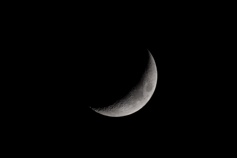 A close-up of a crescent moon, arcing across the bottom-right of the image. Deep craters along the terminator are cast in long shadows, and the lit side is full of details in variegated grey until it turns to white along the very edge.