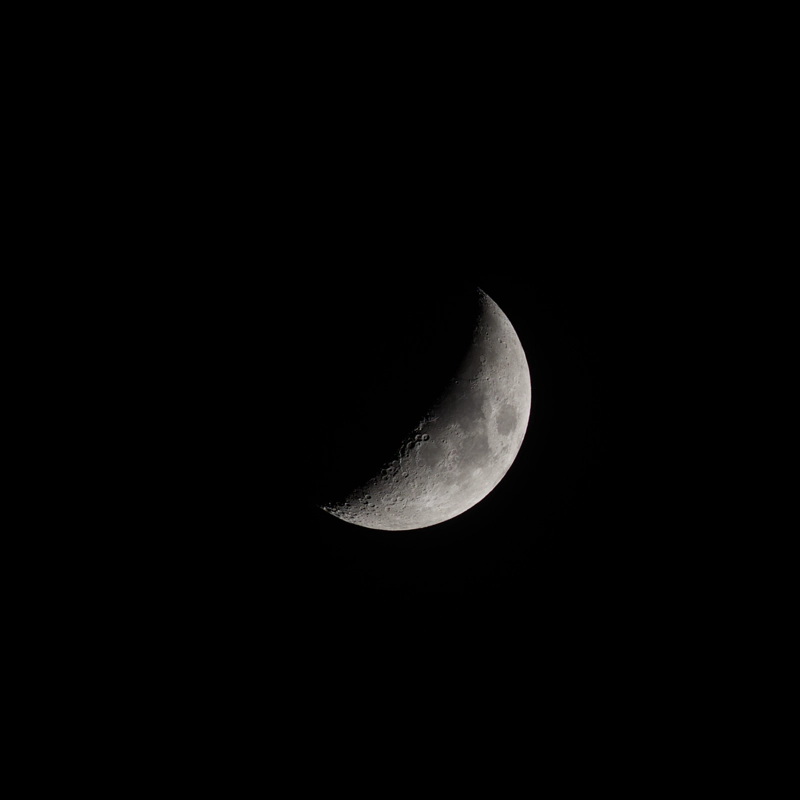 A square image of a wide crescent moon, approaching half-lit, against a black sky. There is a cluster of deep craters near the centre of the image revealed by the changing moon phase.