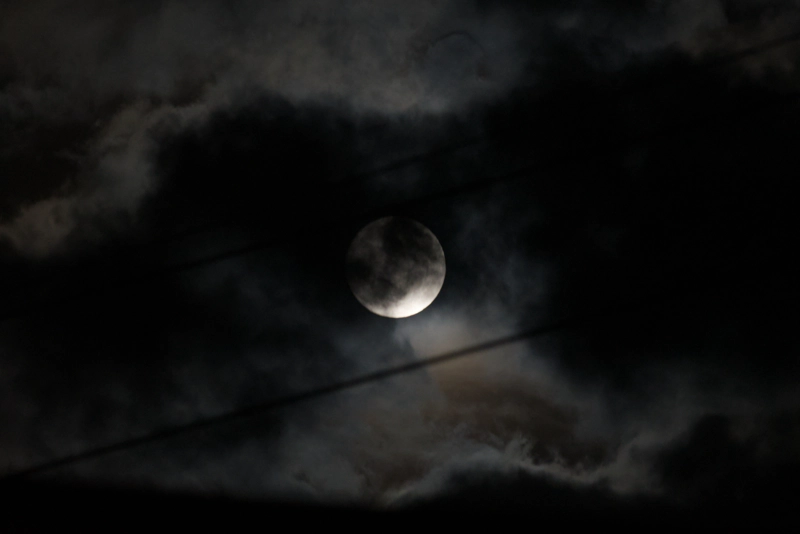 A somewhat-blurry photo of the moon peeking out from behind dense clouds, between power lines. The surrounding cloud is multi-layered, and the moonlight casts fascinating pinks and browns across the different layers.