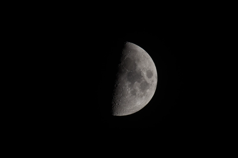A grey half-moon against a black sky. Large deep-grey seas can be seen on the lit half of the moon, and along the terminator, craters cast long shadows until they vanish into the lunar night.