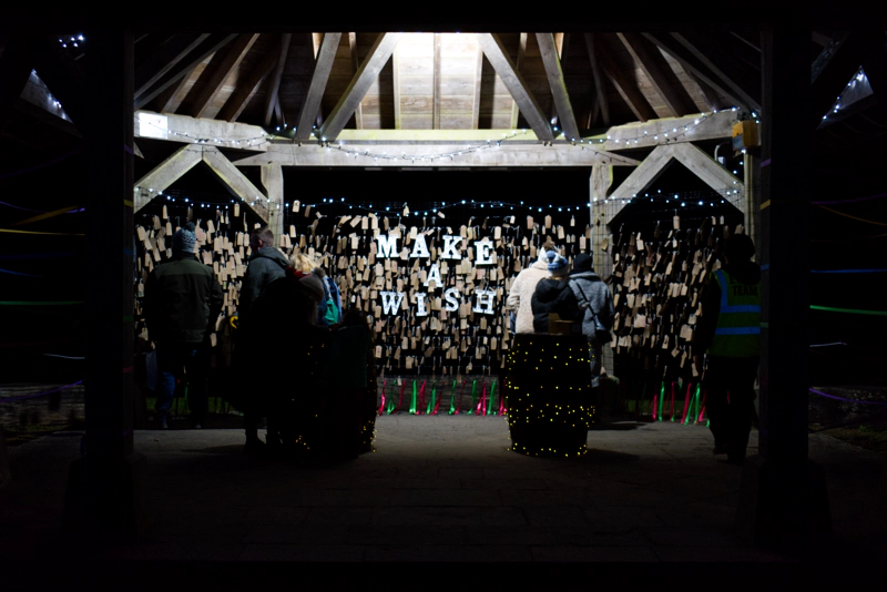 A cluster of people in winter clothes stand in a wooden gazebo, decorated with white fairy-lights. The words "MAKE A WISH" hang in the gazebo, surrounded be hundreds of wishes written on little brown-paper cards.