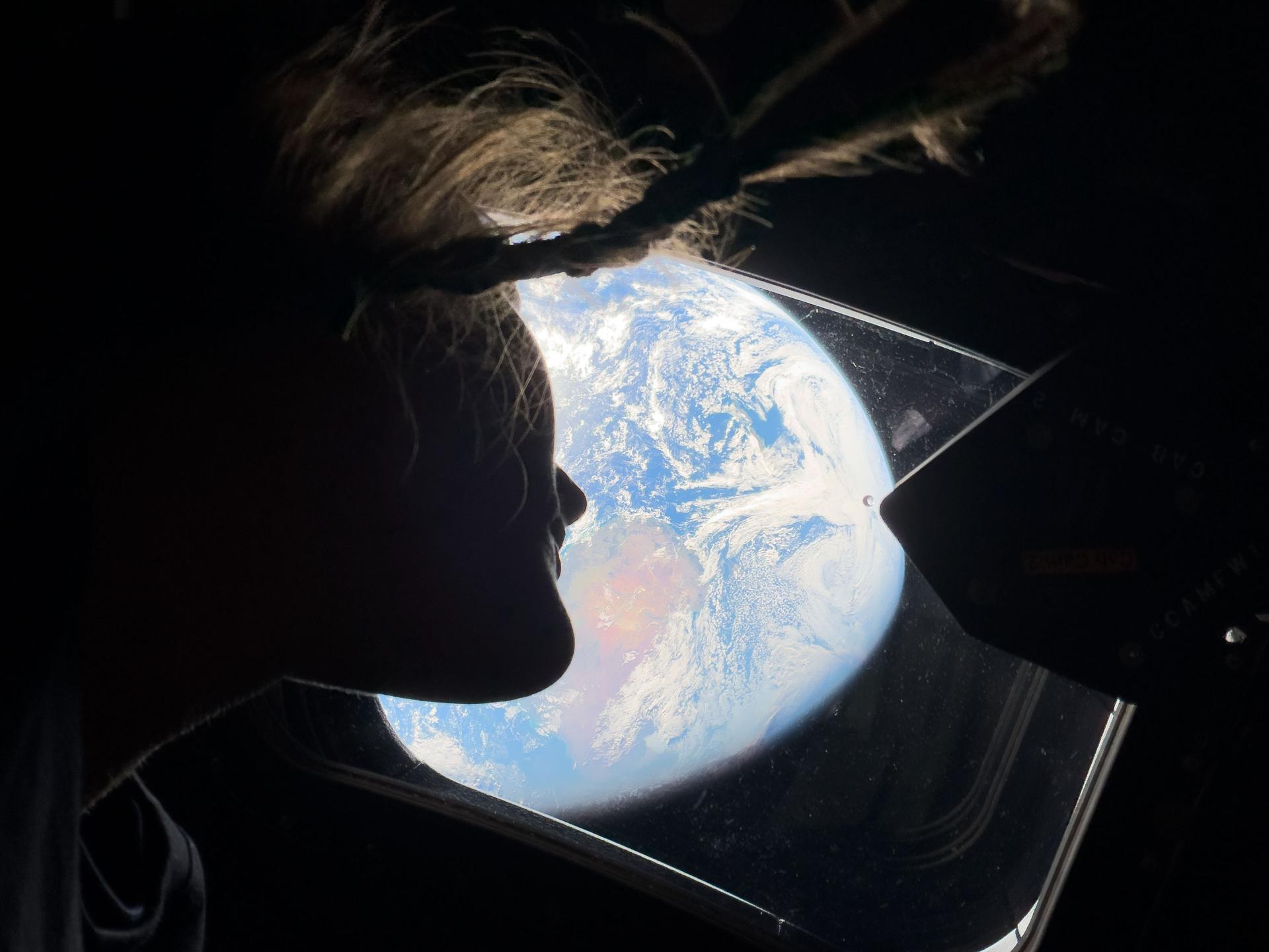 Astronaut Christina Koch looks out through the capsule window at Earth spinning slowly beneath them. Her braided hair wafts in zero gravity, catching the light reflected by Earth.